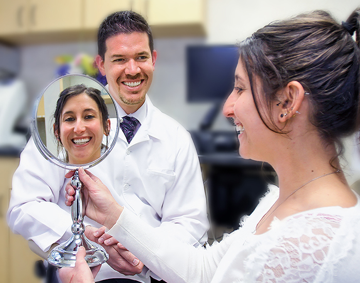 A male dentist is smiling at a female patient who is looking at a mirror checking her smile