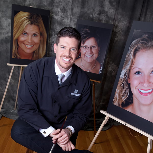 Dr. Nosti Painter of Smiles Dr. Nosti sitting on a chair holding a paintbrush at his background are paintings of a picture of three woman with a gorgeous smile