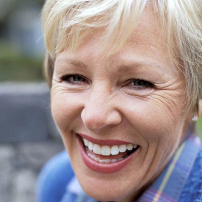 Dentures help restore your smile in a removable and affordable way. An old woman with white teeth and blonde hair smiling and wearing a sweater